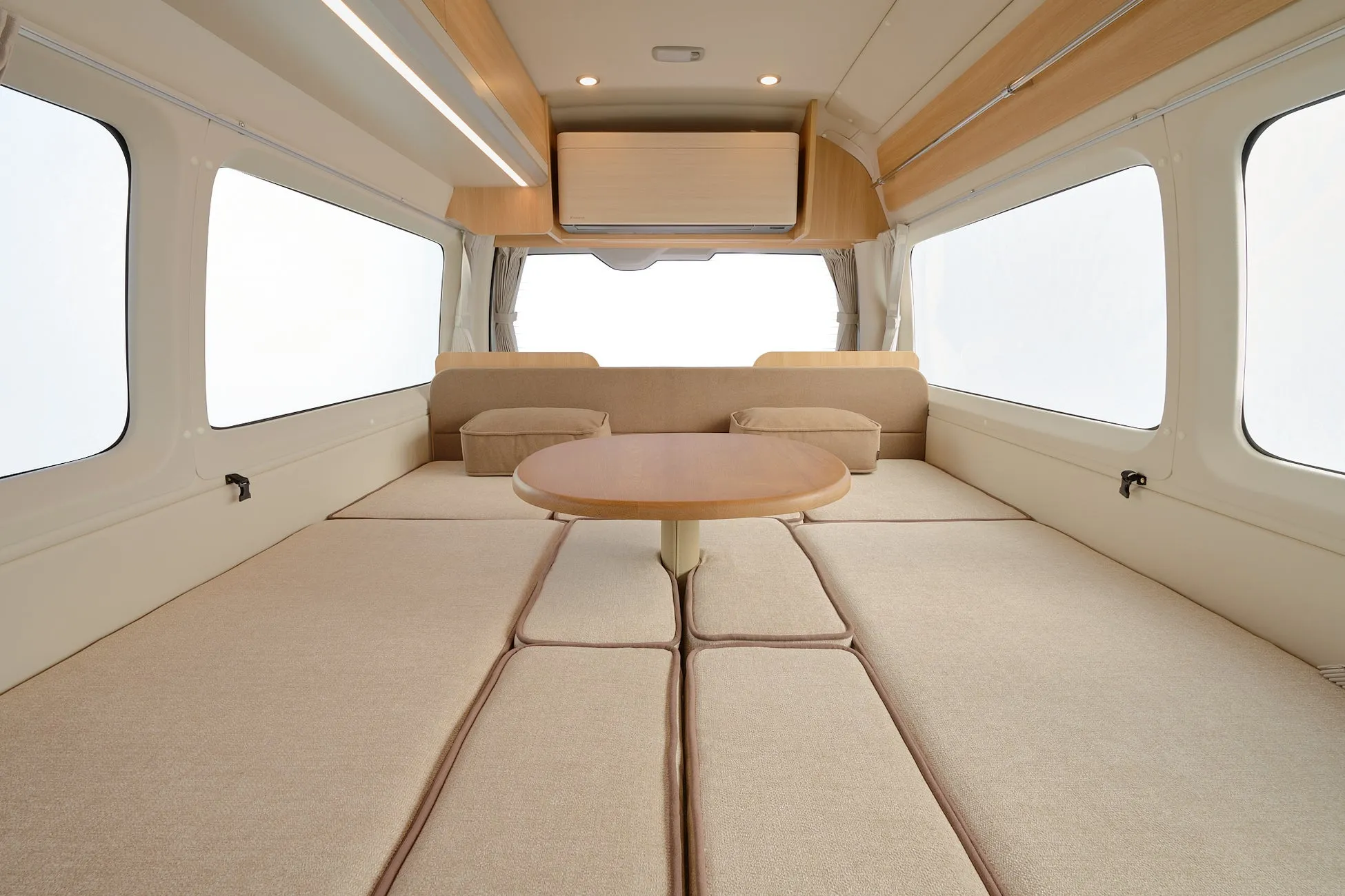 キャンピングカーの内部の様子。中央にテーブルとクッション、両側に窓、ベージュ色の内装。