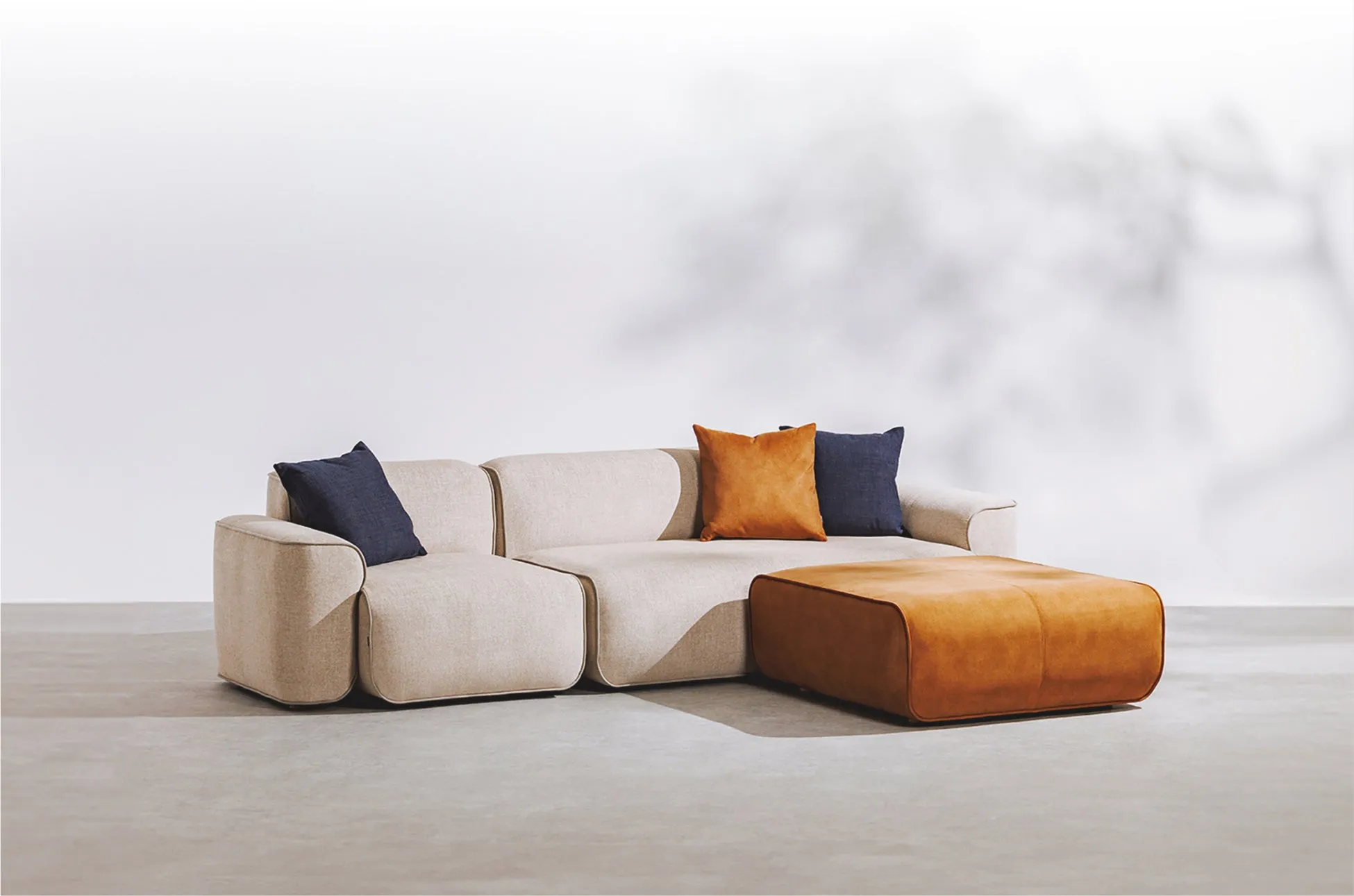 白い壁の前に置かれた、ベージュのソファとオレンジ色のオットマンの画像です。ソファには3つのクッションが置かれており、そのうち2つは濃い青色、もう1つはオレンジ色です。床はコンクリートのようです。