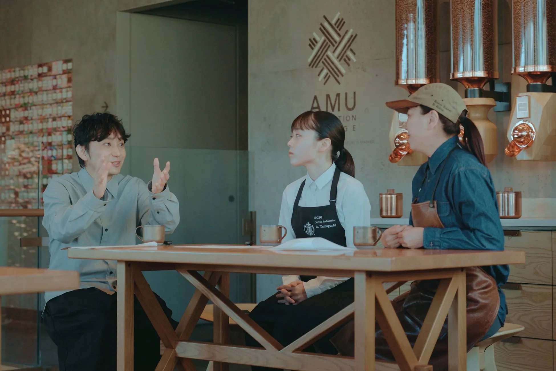 3人の人々がテーブルを囲んで話している様子。中央の女性はエプロンを着けており、左側の男性は両手を広げて話している。背景にはコーヒー豆の容器やロゴが見える。