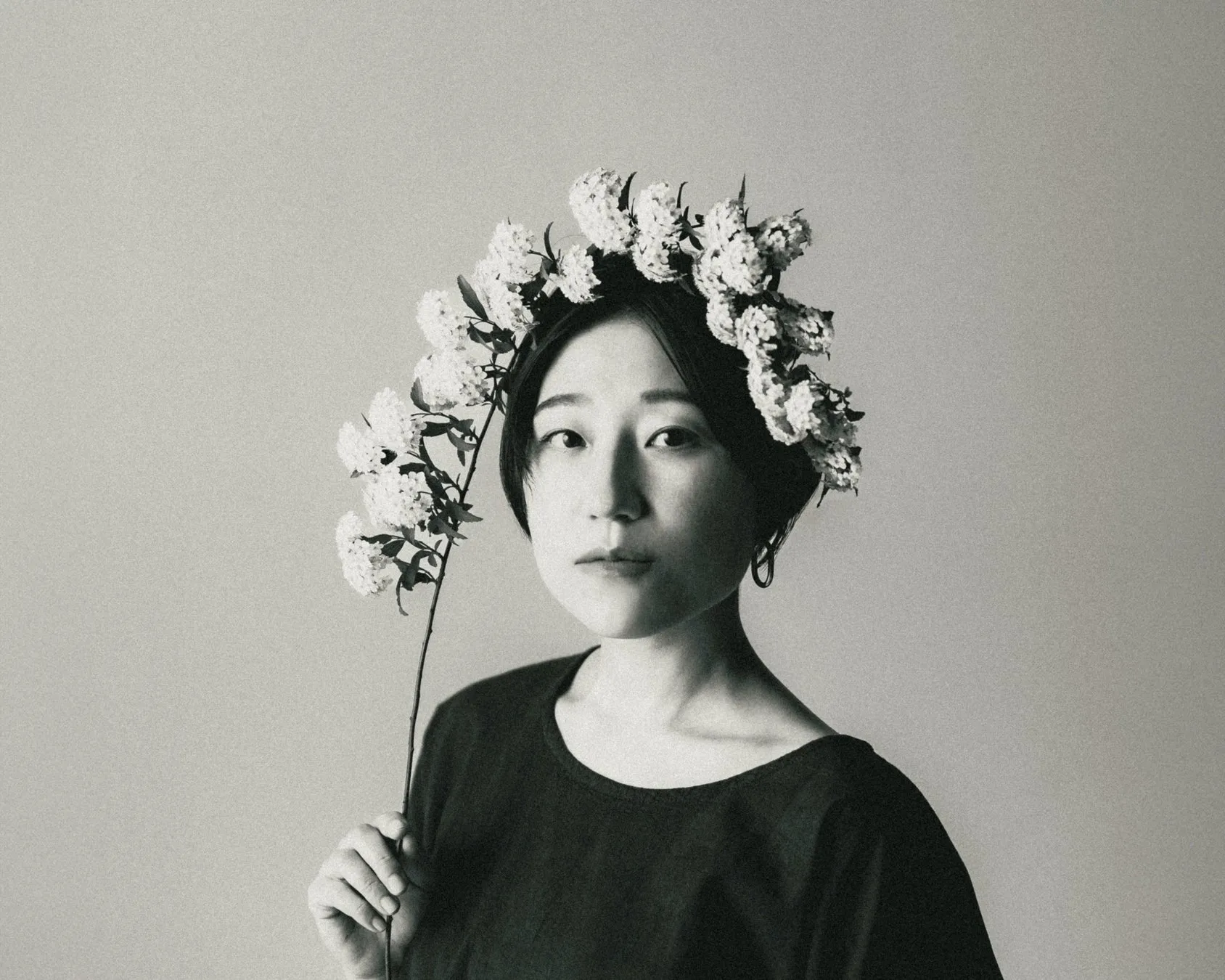 花冠をつけた女性のモノクロポートレート。女性は、白い花が咲いた枝を持ち、カメラ目線で少し悲しげな表情をしています。