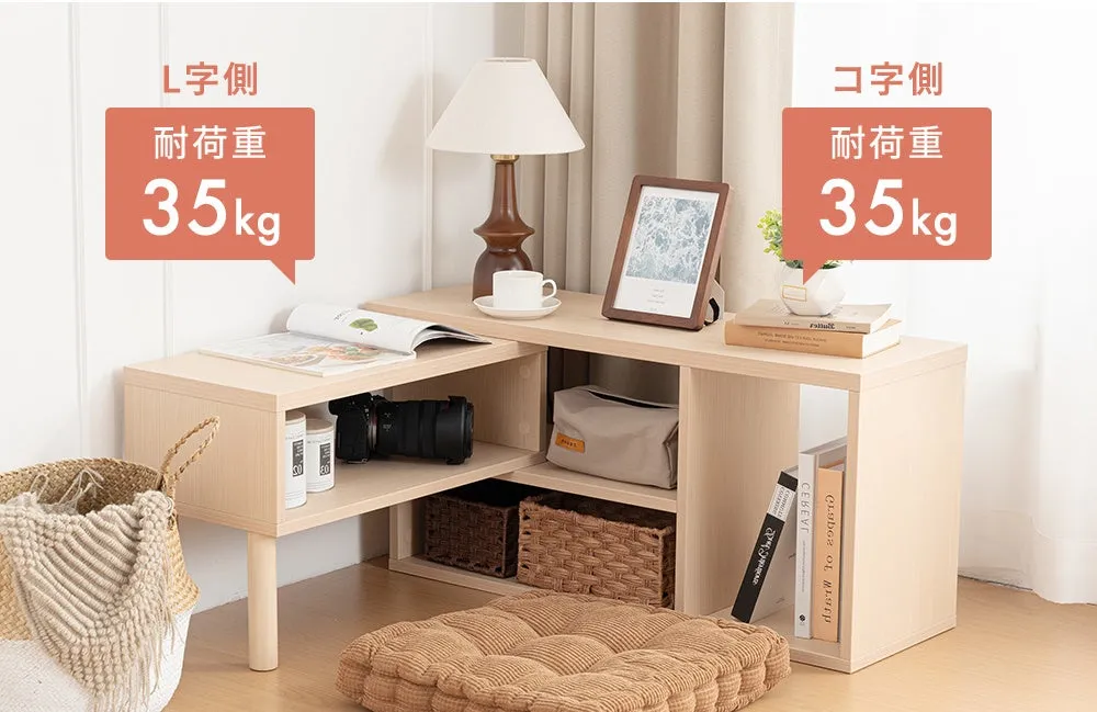 L字型の木製ローテーブルの写真。テーブルの上には、ランプ、写真立て、本、カメラ、装飾品が置かれています。テーブルの下には、バスケットやクッションがあります。