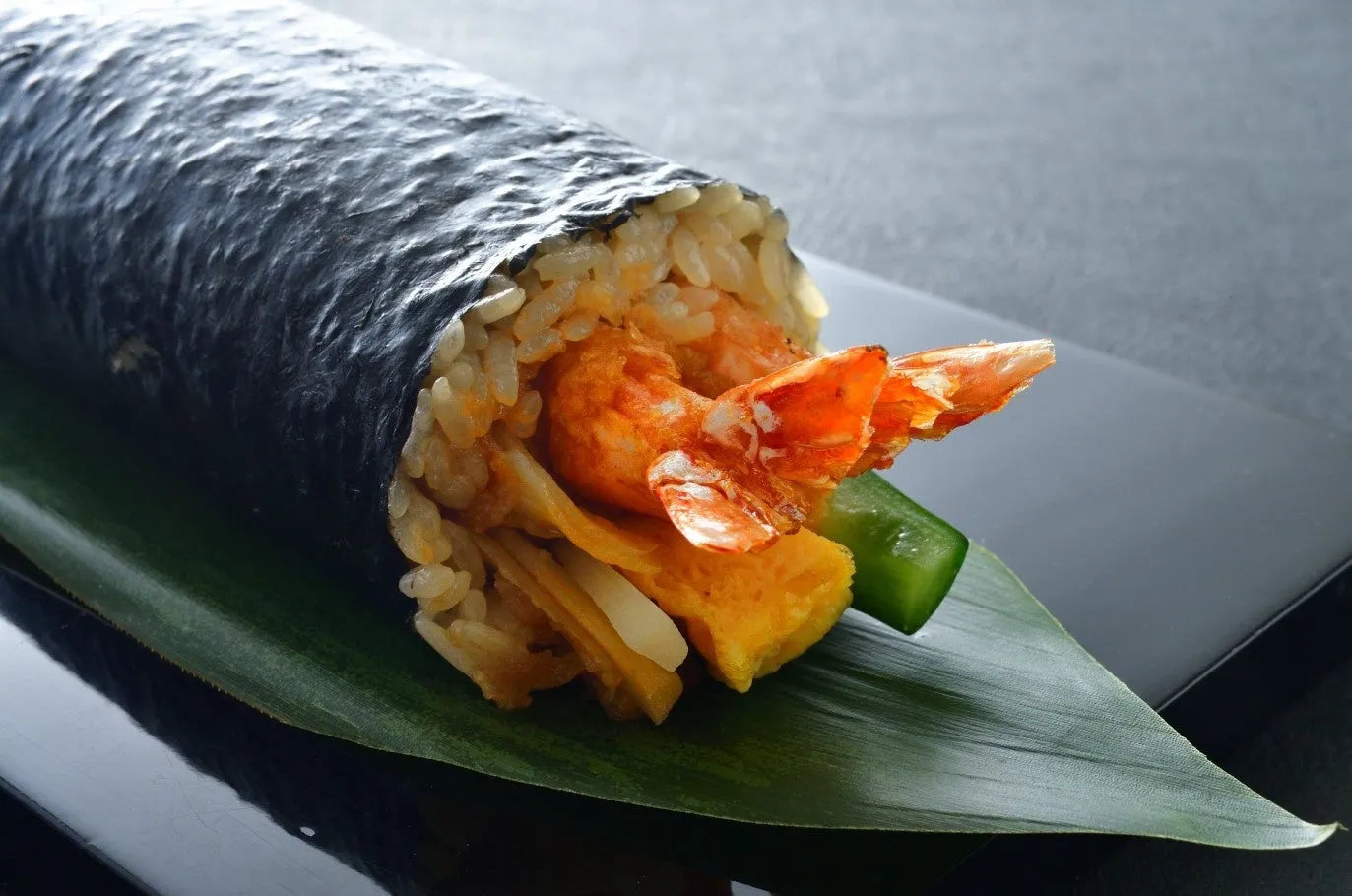 エビ天巻き寿司のクローズアップ写真。エビ天、卵焼き、キュウリ、ご飯が海苔で巻かれている。皿の上に葉が敷かれている。