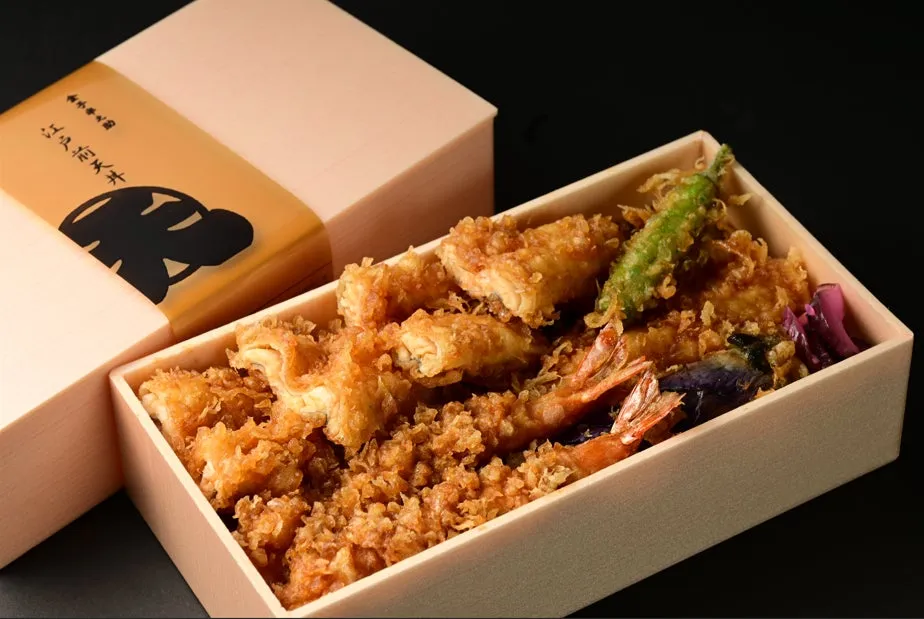 天ぷら弁当の画像です。エビ、魚、野菜の天ぷらが詰められており、弁当箱の蓋には文字とイラストが印刷されています。