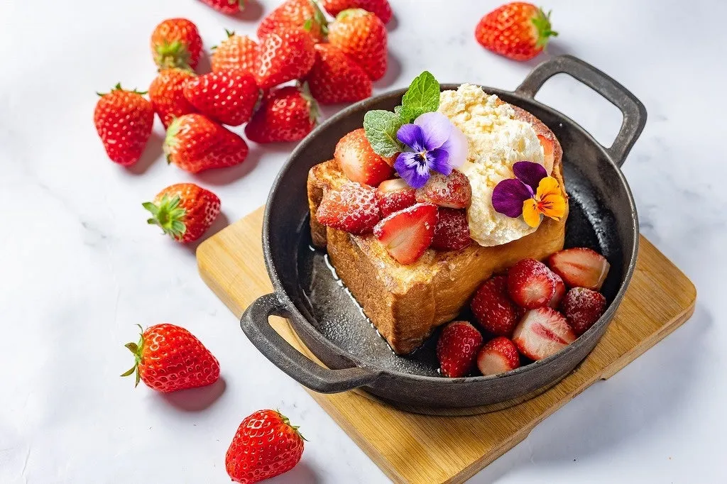 いちごが添えられたフレンチトーストの画像です。フレンチトーストは黒い鉄製の器に盛り付けられており、ホイップクリームとエディブルフラワーがトッピングされています。周りには、たくさんの新鮮ないちごが散りばめられています。