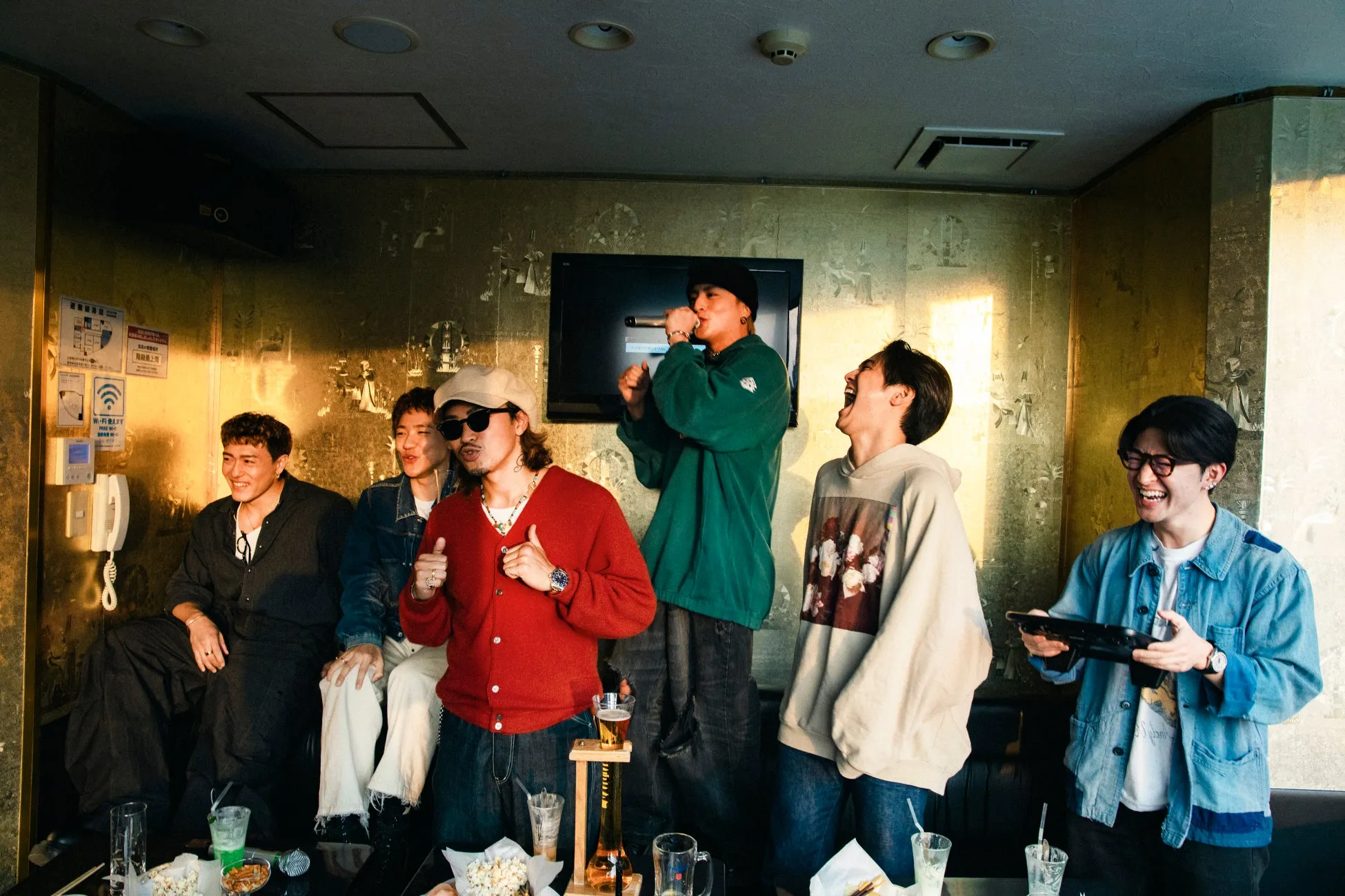 カラオケボックスで楽しそうに歌う男性グループの写真。それぞれ異なるファッションに身を包み、マイクを握ったり、笑ったり、思い思いの時間を過ごしている様子が写っています。