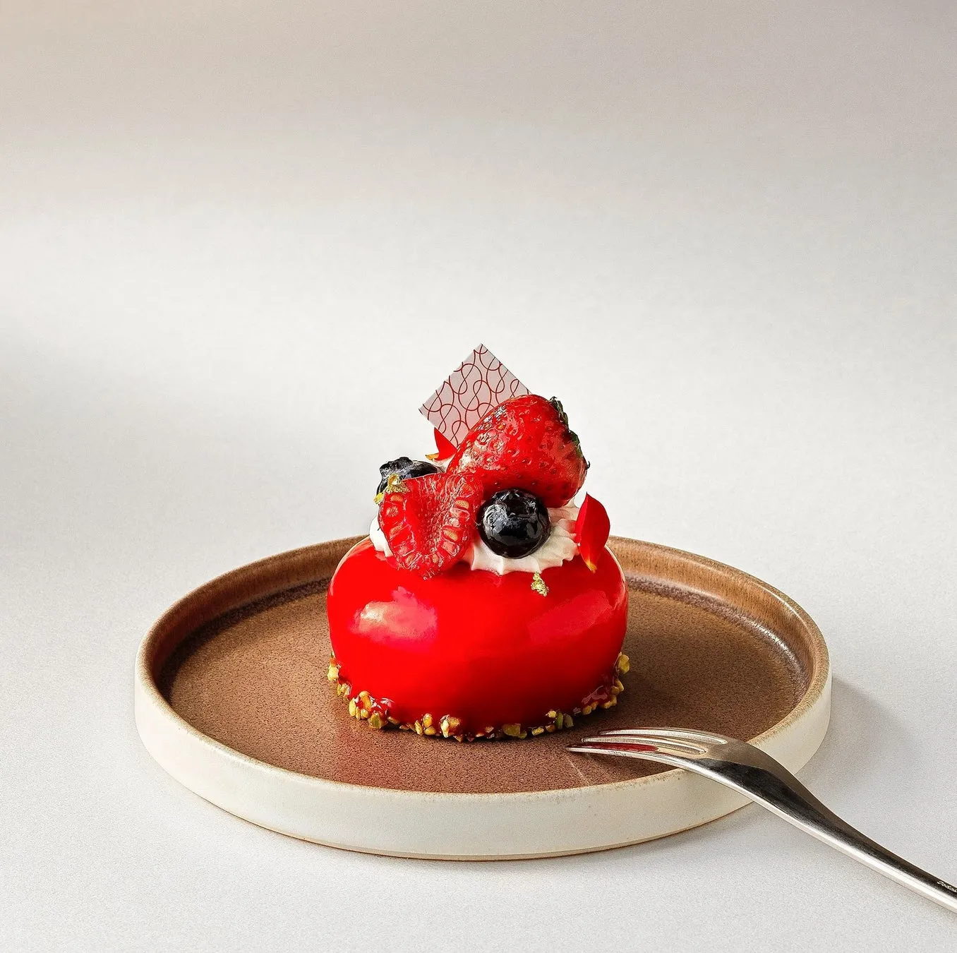 赤く艶やかなケーキが皿に載せられており、その上にベリー類が飾られている。フォークが添えられている。