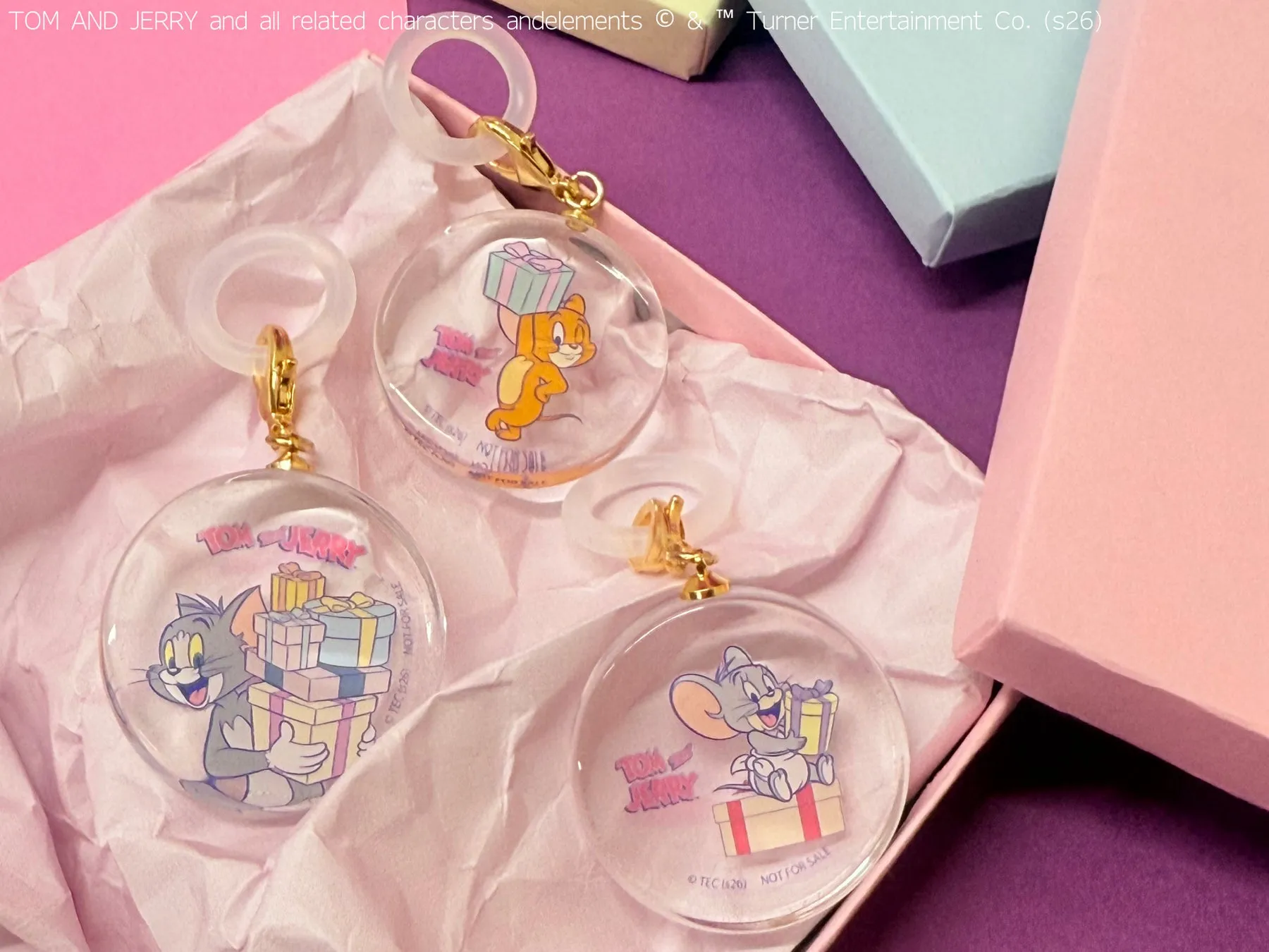 トムとジェリーのキャラクターが描かれたキーホルダーの写真です。キーホルダーは、プレゼントを持ったトムとジェリーが描かれており、ピンクのギフトボックスと紙に囲まれています。