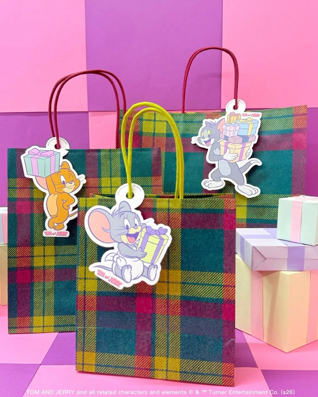 トムとジェリーのキャラクターが描かれたギフトバッグの写真。ギフトバッグはチェック柄で、トムとジェリーのキャラクターが描かれたタグが付いています。背景には、ピンクと紫の壁と、いくつかのプレゼントがあります。