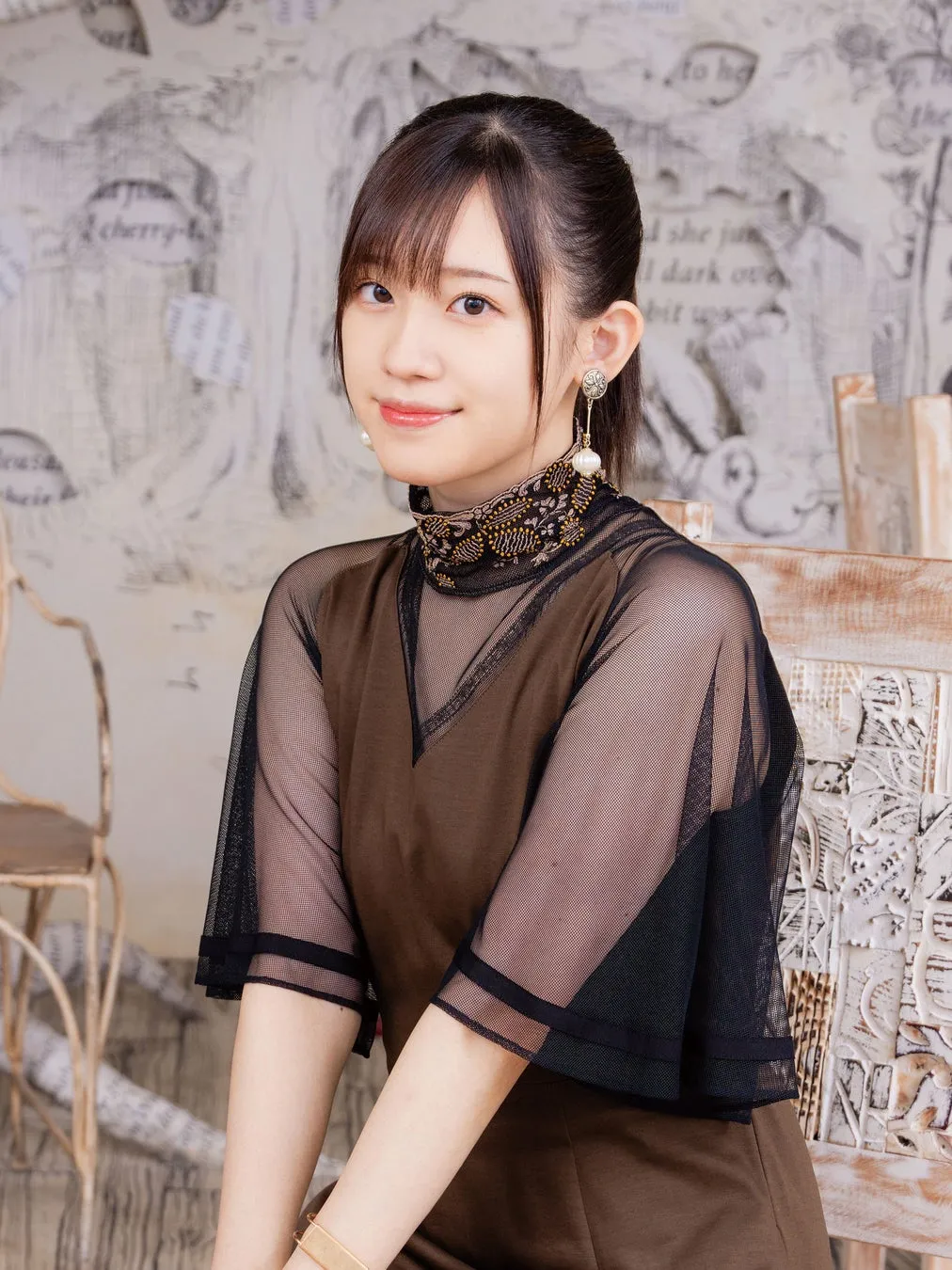 女性が椅子に座ってカメラ目線で微笑んでいる写真。彼女は黒いメッシュのトップスと茶色のドレスを着ており、髪は後ろでまとめています。背景は、テキストとパターンで装飾された壁です。