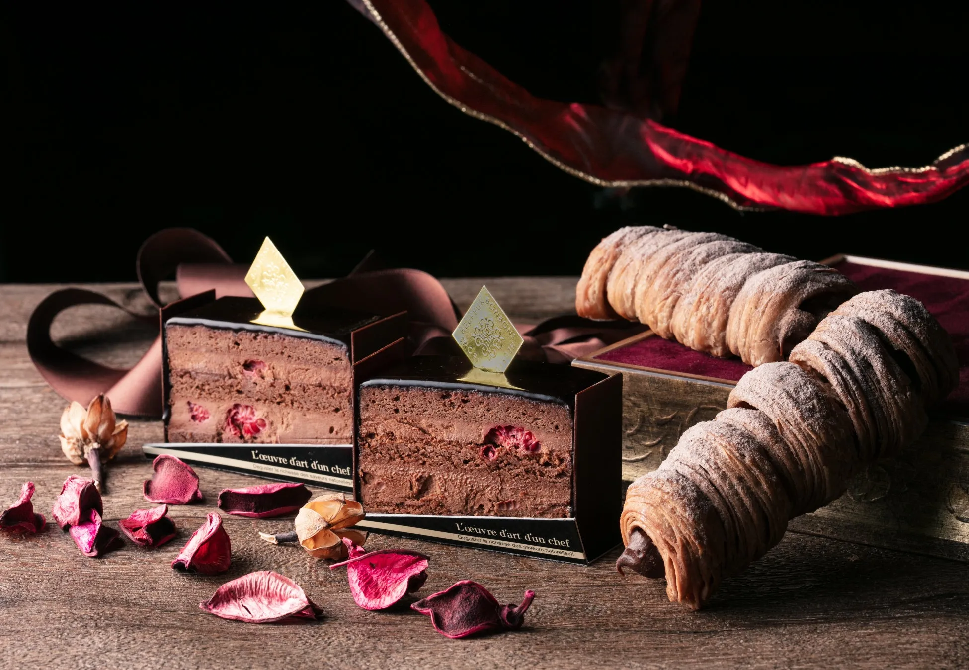 チョコレートケーキとクロワッサンが置かれた写真。ケーキはチョコレートでコーティングされ、金色の飾りが施されている。クロワッサンは粉砂糖がまぶされており、背景には赤い布と木製のテーブルが写っている。
