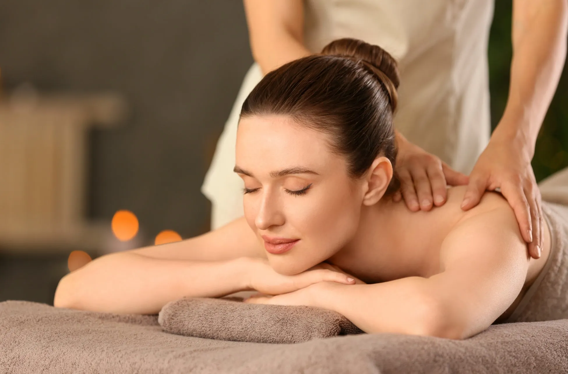 女性がマッサージを受けている様子。彼女は目を閉じ、リラックスした表情をしている。セラピストの手が彼女の肩に置かれ、マッサージをしている。