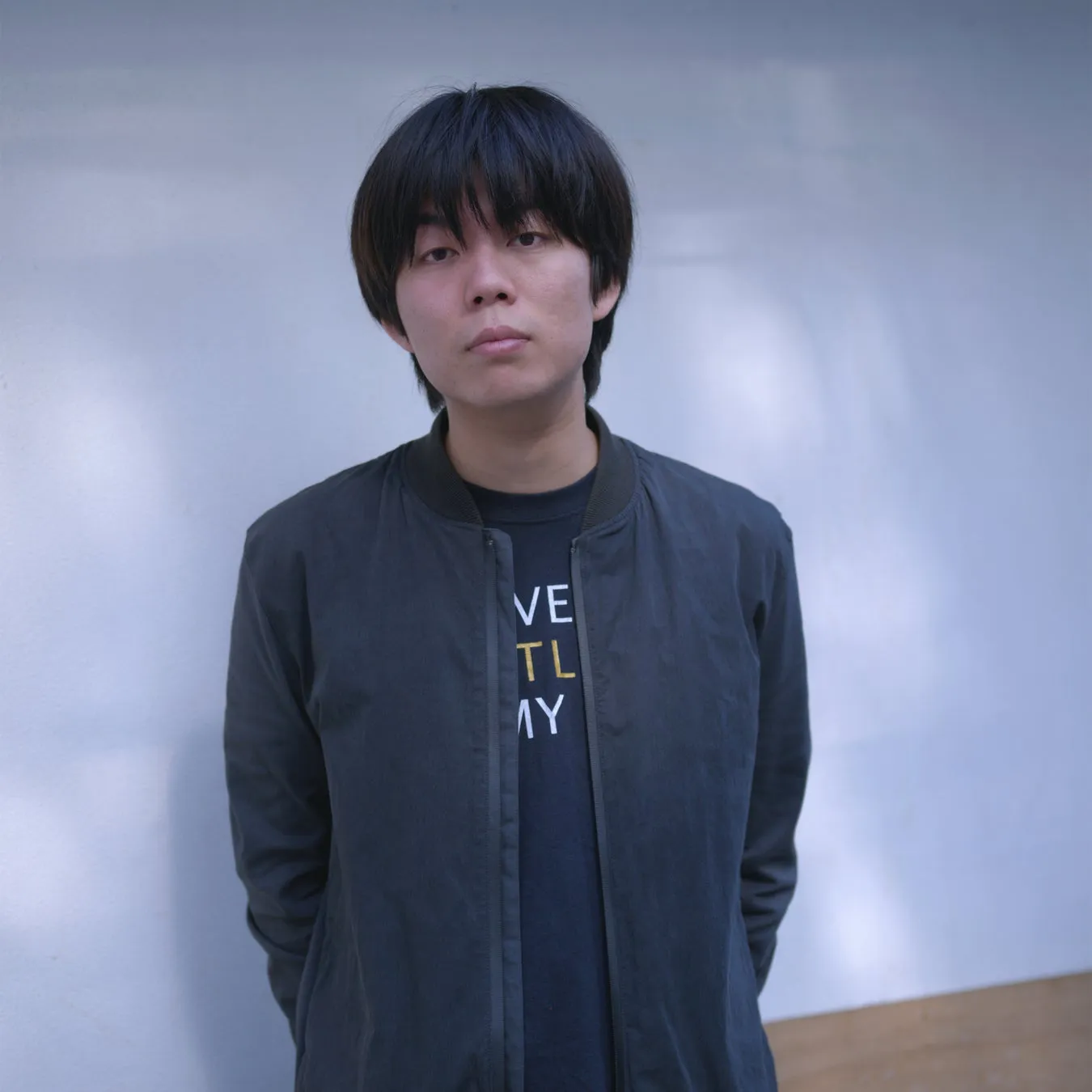 壁を背景に、黒髪の男性がカメラ目線で立っている写真