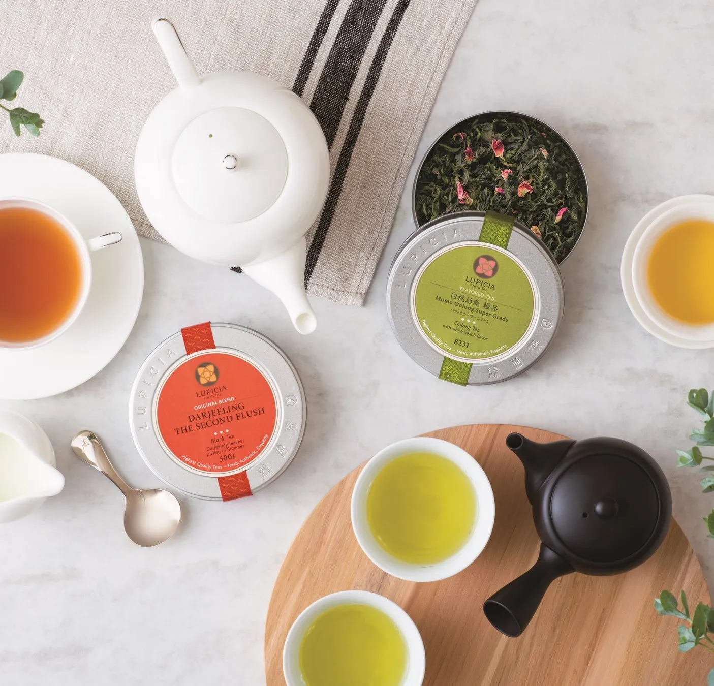 様々な種類のお茶のセットの写真。白のティーポットとティーカップ、茶葉の缶、お茶が入ったカップが写っています。お茶の種類は、ダージリン、白桃烏龍茶などです。