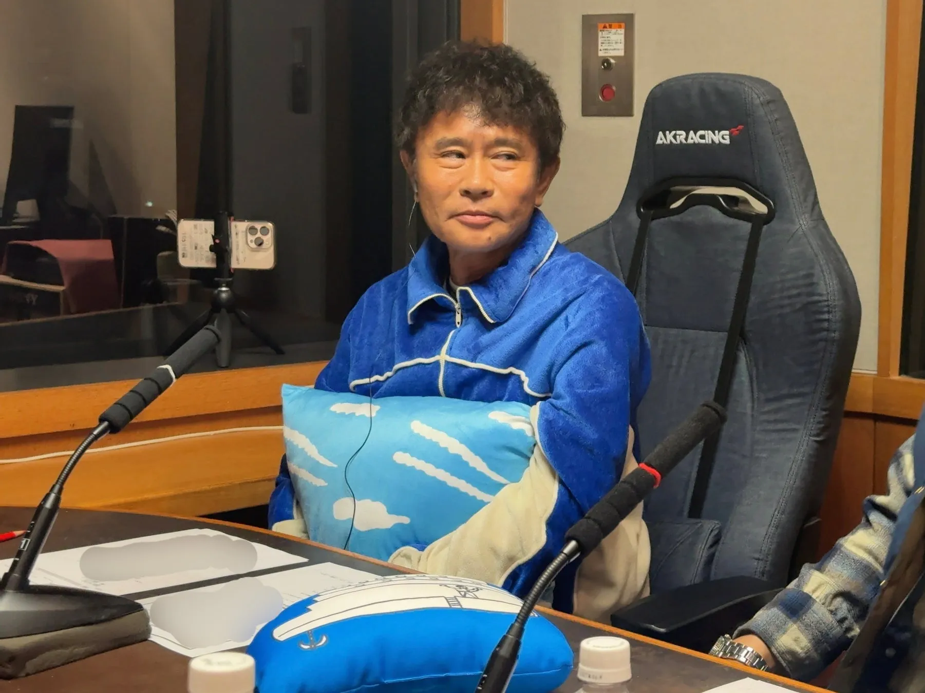 男性が青い服を着て、クッションを抱えながら、マイクの前で座っています。背景には、AKRACINGの椅子があります。