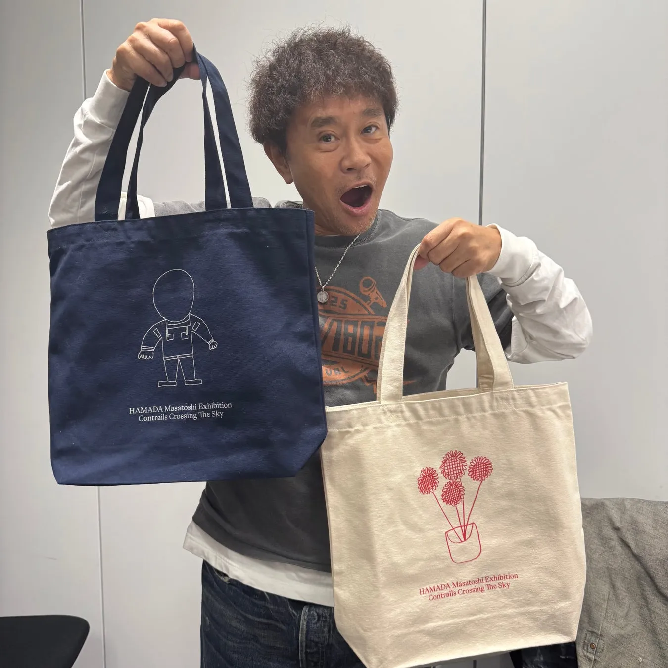浜田雅功がトートバッグを2つ持って笑顔で写っている写真。片方のバッグには宇宙飛行士のイラストが、もう一方には花のイラストが描かれている。