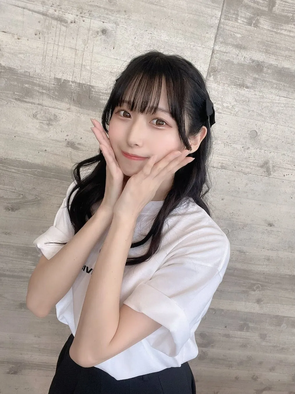 女性が両手で頬に触れて笑顔を見せている写真です。彼女は黒髪で、白いTシャツと黒いスカートを着ています。背景は灰色の壁です。
