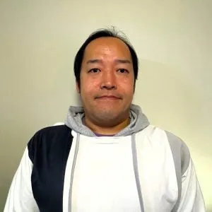 男性が正面を向いて立っている姿を捉えたポートレート写真です。男性は、白、グレー、紺色のフード付きのパーカーを着ています。背景は薄いベージュ色です。