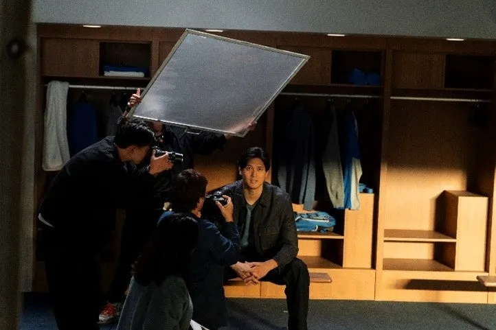 更衣室で撮影されている男性の写真。周囲にはカメラマンや照明機材があり、撮影の様子が伺える。