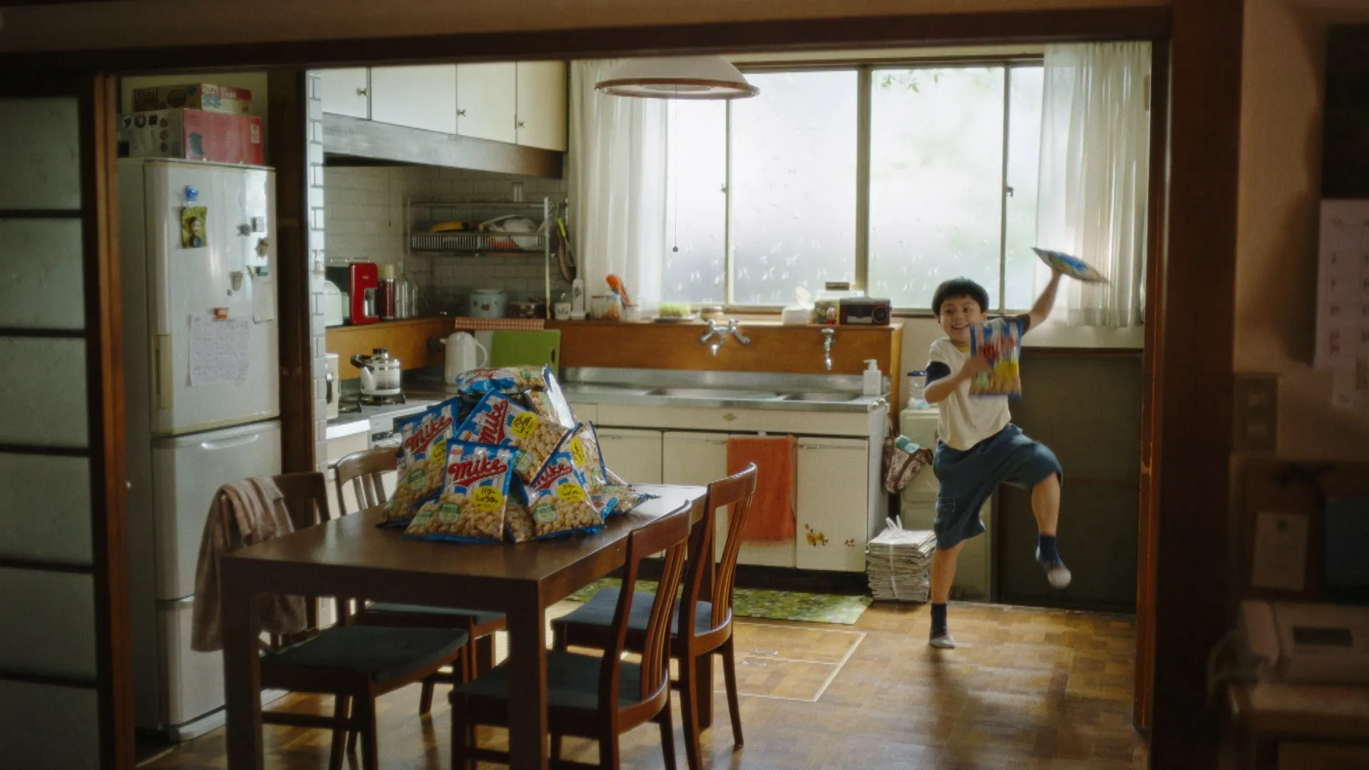 キッチンで皿を掲げて喜ぶ少年。テーブルには大量のポテトチップスが置かれている。