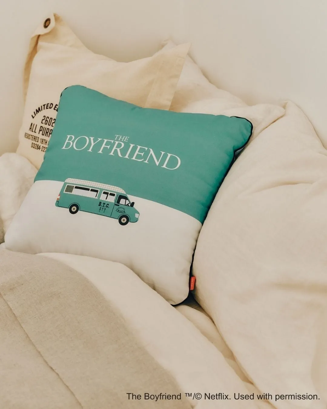 枕の上に「THE BOYFRIEND」の文字とバスのイラストが描かれた枕が置かれている写真です。背景には、他の枕や寝具が見えます。
