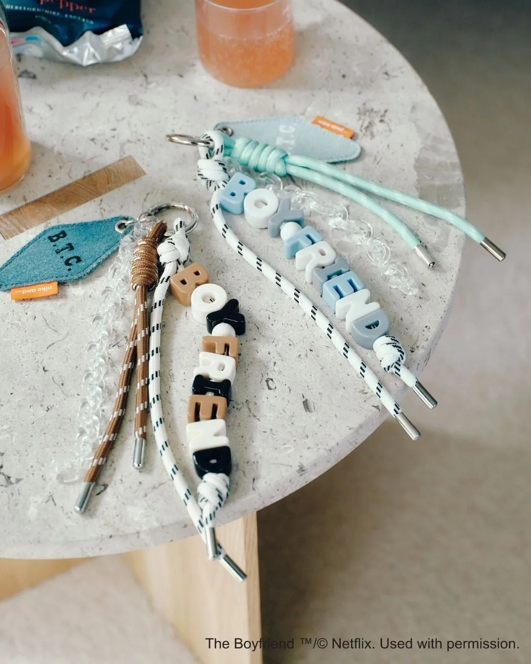 テーブルの上に、文字が書かれたキーホルダーが複数置かれている写真。キーホルダーは、ロープや紐で構成され、様々な色やデザインの文字が組み合わされている。背景には、飲み物やその他の小物も見える。