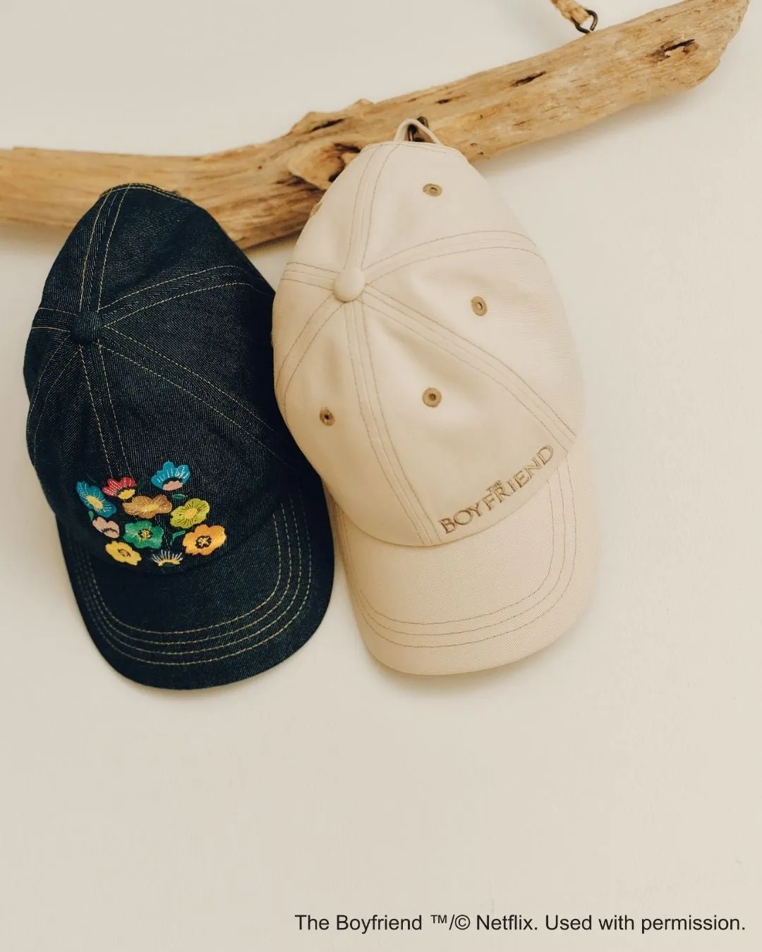 2つの野球帽が木製の枝に掛けられており、片方は花柄の刺繍、もう片方は「BOYFRIEND」の文字が刺繍されています。