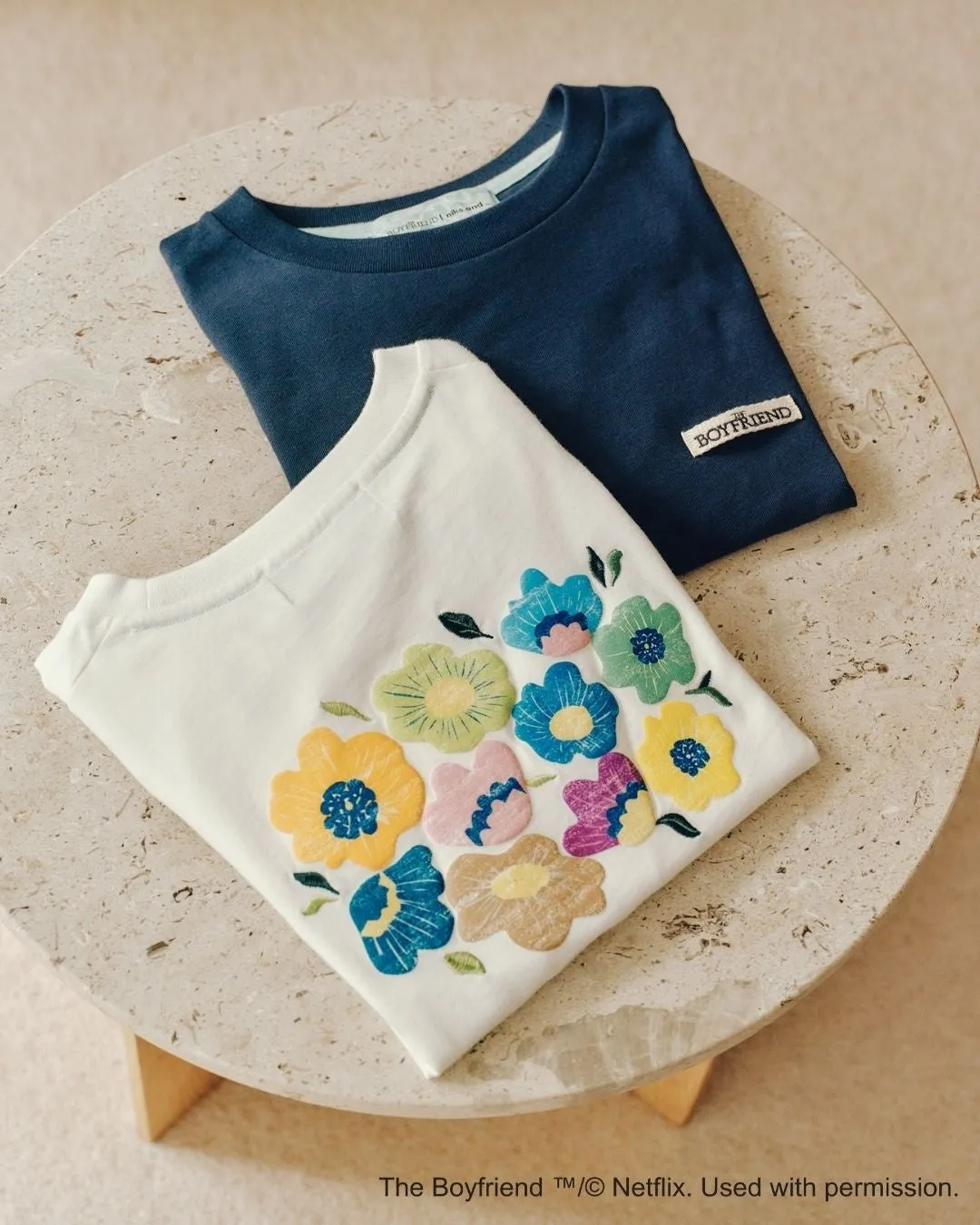 白いTシャツと濃い青色のTシャツが、丸いテーブルの上に重ねて置かれています。白いTシャツには、様々な色の花の刺繍が施されています。青いTシャツには、ブランド名が記載された小さなタグが付いています。
