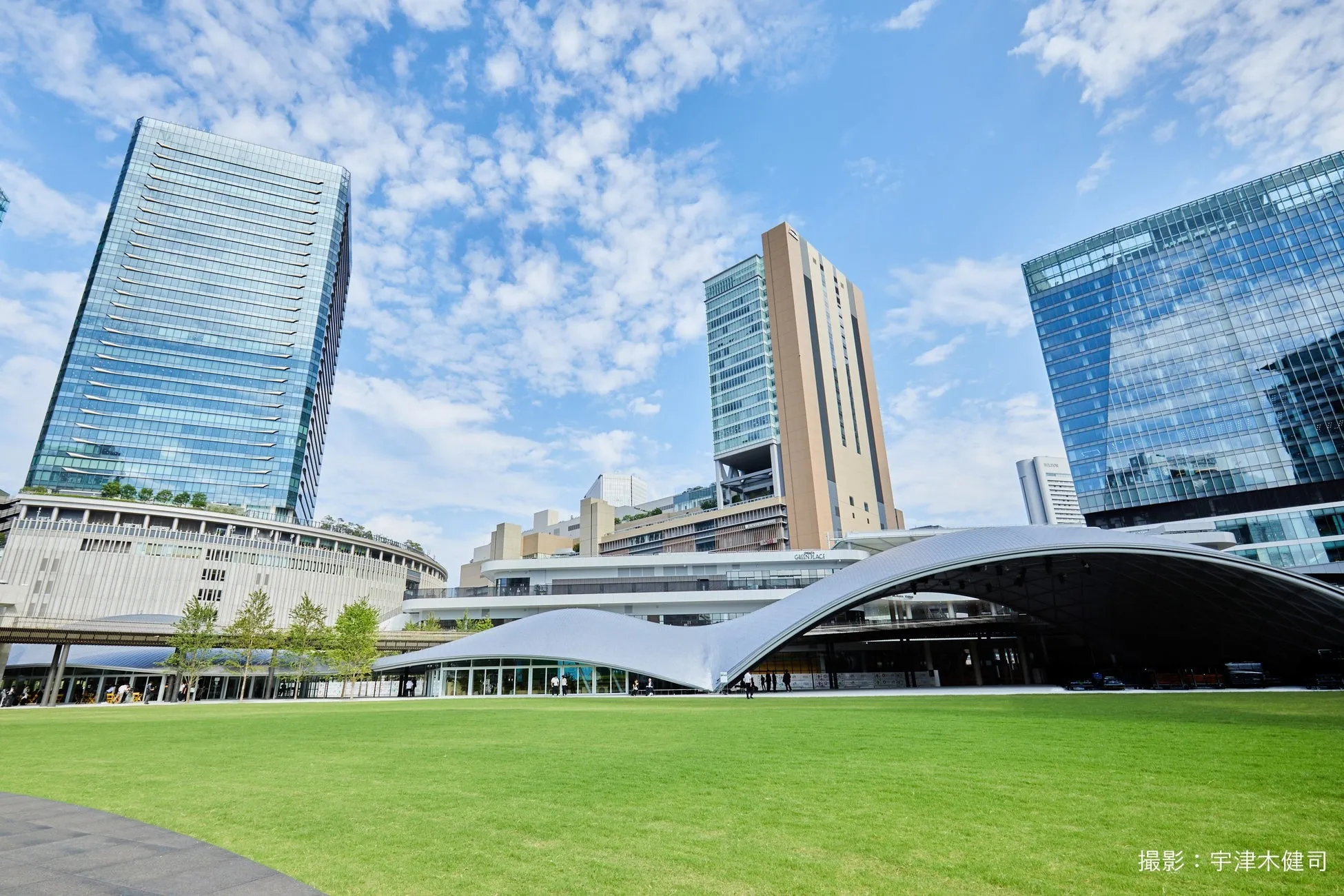 青空の下、緑豊かな広場と近代的な建物が立ち並ぶ風景写真。ガラス張りの高層ビルや特徴的なデザインの屋根が印象的。