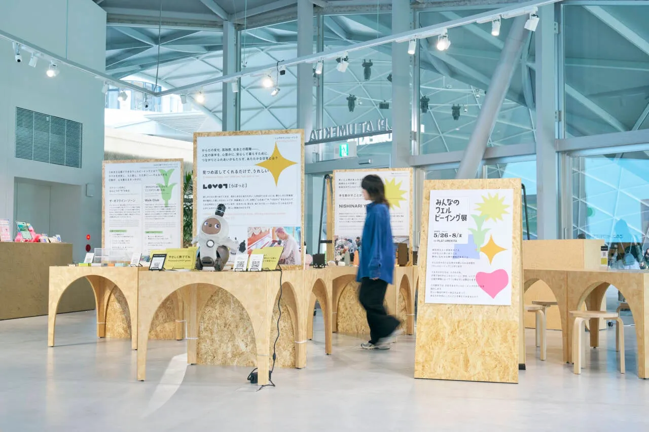 展示会の会場の様子。木製のテーブルに展示物が並び、ロボットやポスターが配置されている。女性が歩いている様子も写っている。