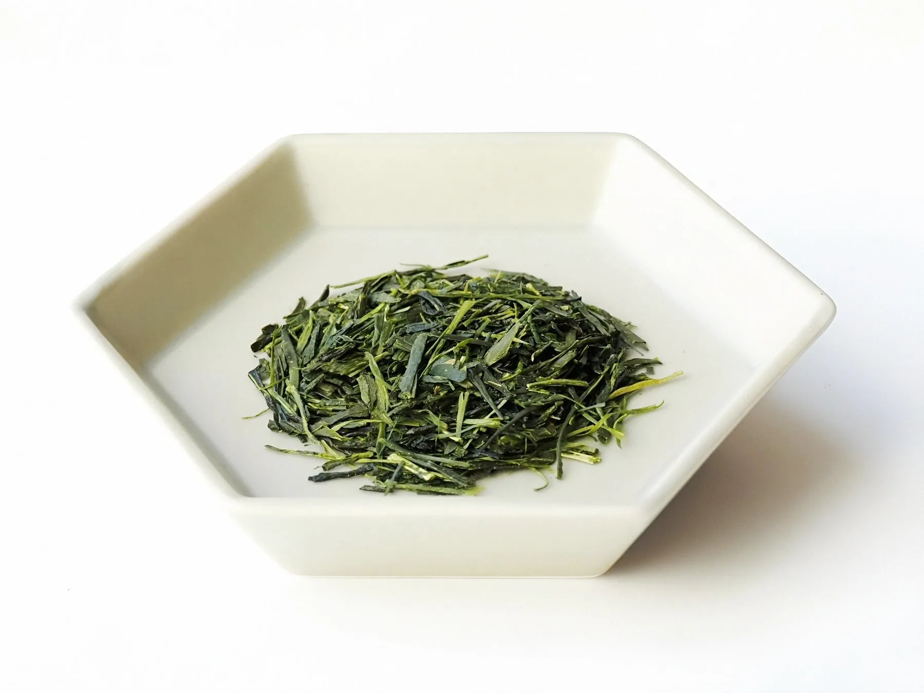 六角形の白い器に入った緑茶の葉のクローズアップ写真。葉は細かく刻まれており、鮮やかな緑色をしている。