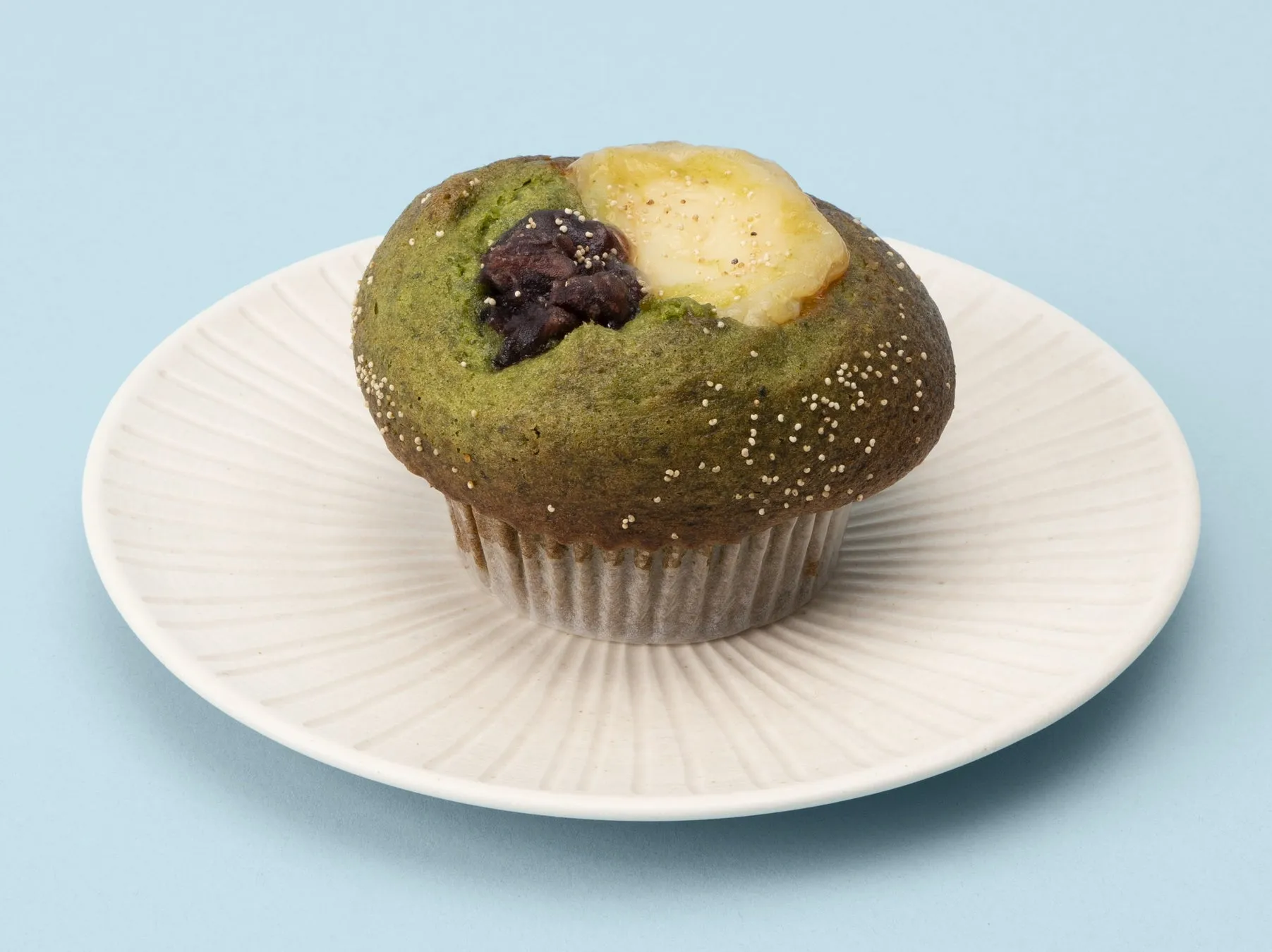 抹茶マフィンが白い皿の上に置かれています。マフィンの上には、小豆と何か白いものが乗っています。背景は水色です。