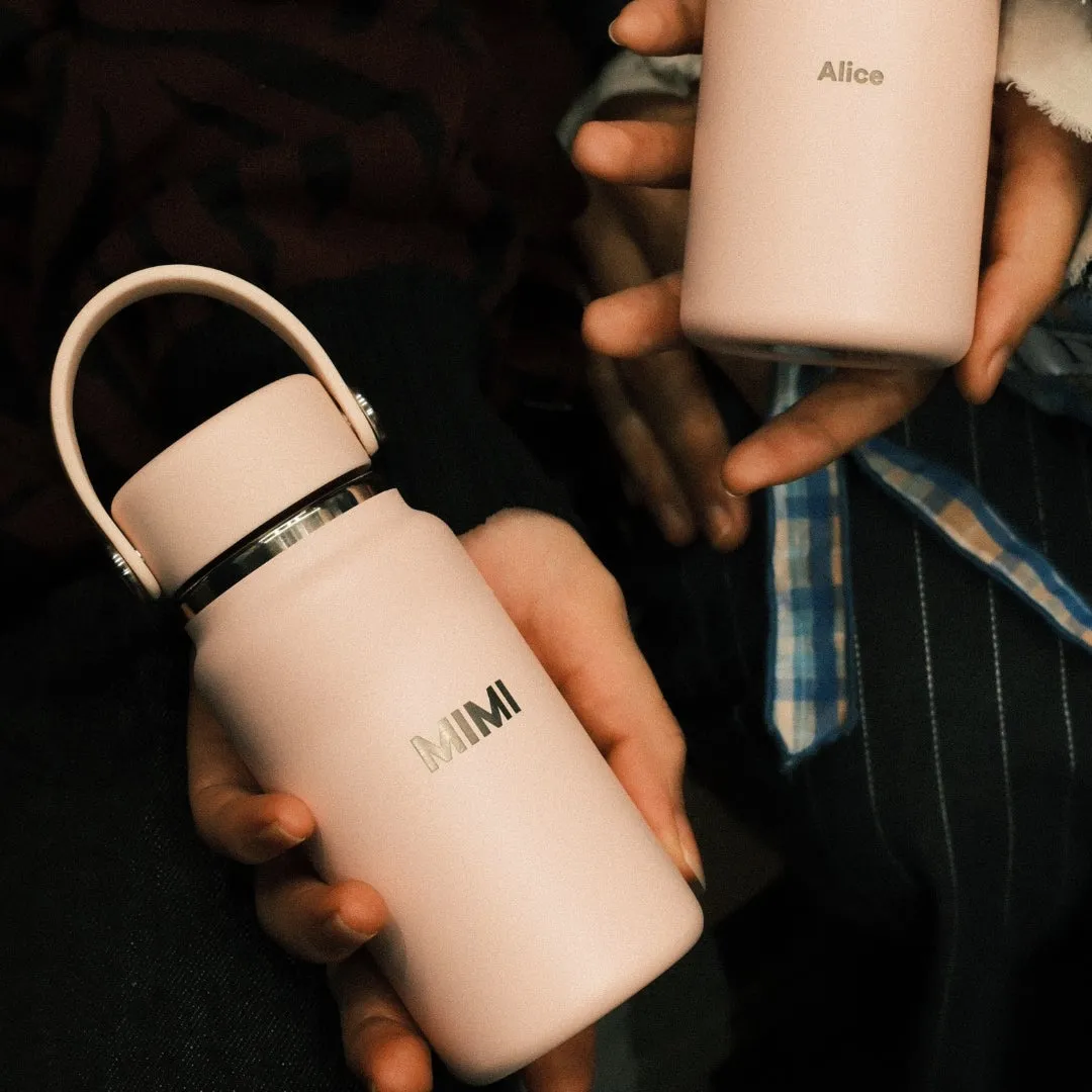 2つのピンク色の水筒を手に持っている様子。片方の水筒には「MIMI」の文字、もう一方には「Alice」の文字が書かれています。