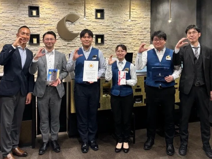 6人の人々が、店の前で写真撮影のためにポーズをとっています。彼らは皆、笑顔で、親指と人差し指で丸を作っています。店は宝石店のように見え、背景には装飾的な壁があります。