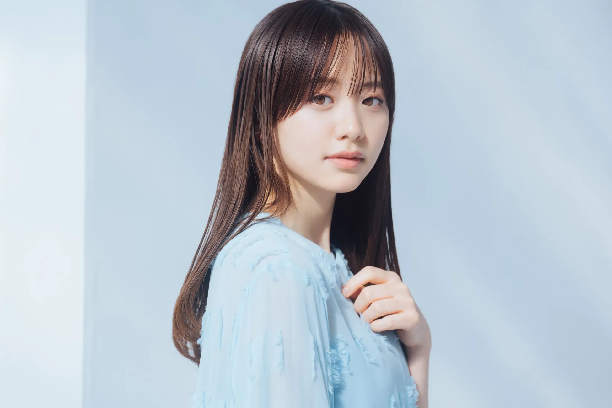 女性のポートレート写真。彼女は長い茶色の髪をしており、薄い青色の服を着ています。彼女はカメラを見つめており、背景は白です。