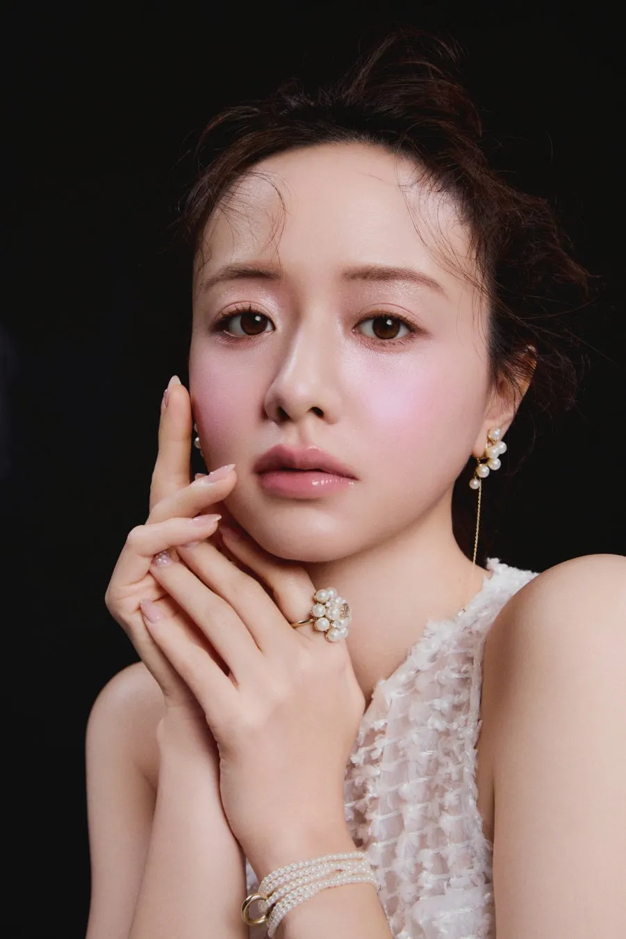 女性のポートレート。白いドレスを着ており、パール風のイヤリング、指輪、ブレスレットを着けている。顔に手を添えている。