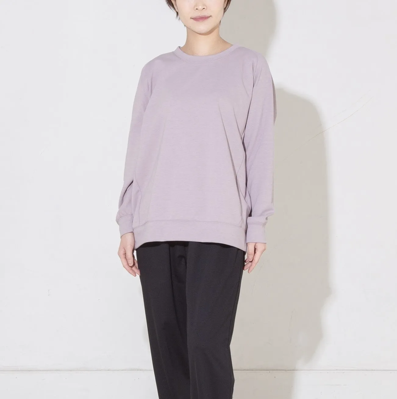 女性が、ラベンダー色の長袖スウェットと黒いパンツを着て立っている写真です。