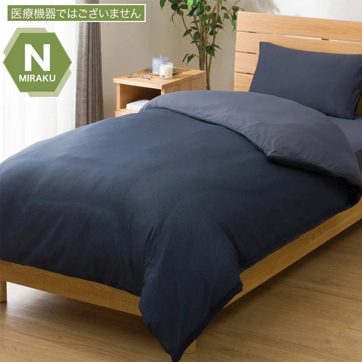 木製のベッドの上に紺色の布団が敷かれた写真です。枕と布団カバーの色が異なり、おしゃれな雰囲気を演出しています。背景には観葉植物やキャンドルが置かれたサイドテーブルがあり、落ち着いた空間を醸し出しています。