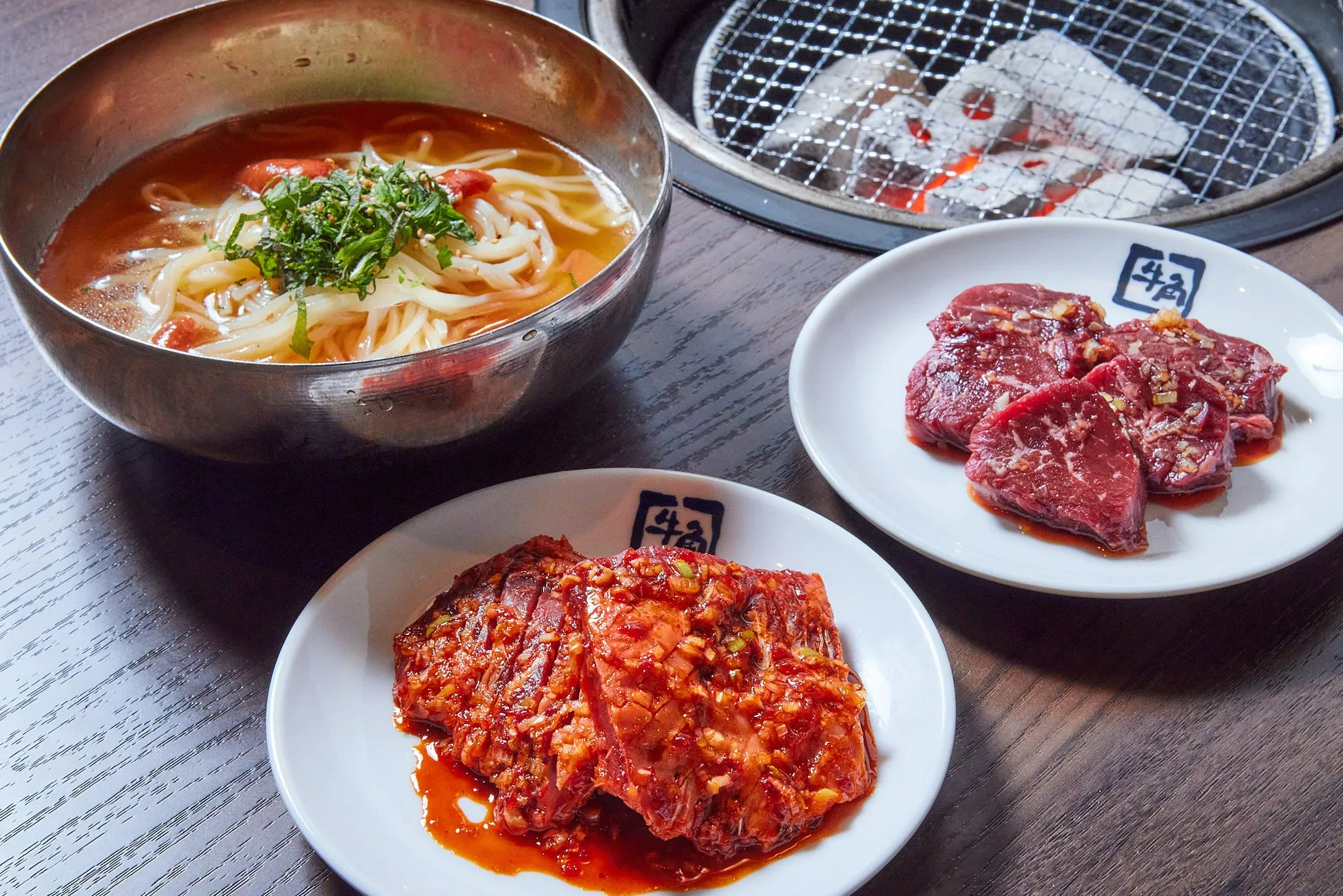 焼肉と麺料理が並んだ食卓の写真です。牛肉は、白い皿に盛り付けられ、タレに漬けられたものと、そのままのものがあります。麺は、金属製の器に入っており、スープが注がれています。炭火焼のコンロもあり、本格的な焼肉店のような雰囲気です。