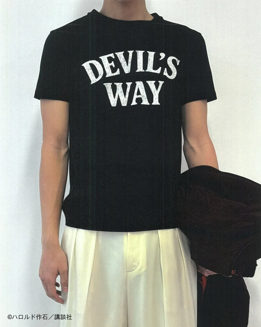 黒いTシャツに「DEVIL'S WAY」と書かれた男性が写っている。白いパンツを履き、ジャケットを持っている。