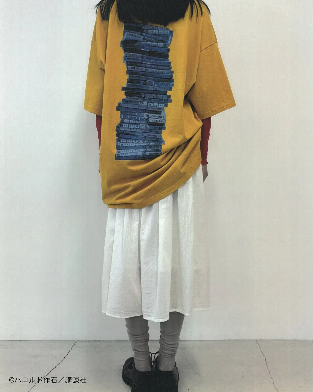 黄色いTシャツに本がプリントされた女性の後ろ姿。白いスカート、グレーのレギンス、黒い靴を履いている。背景は白い壁。