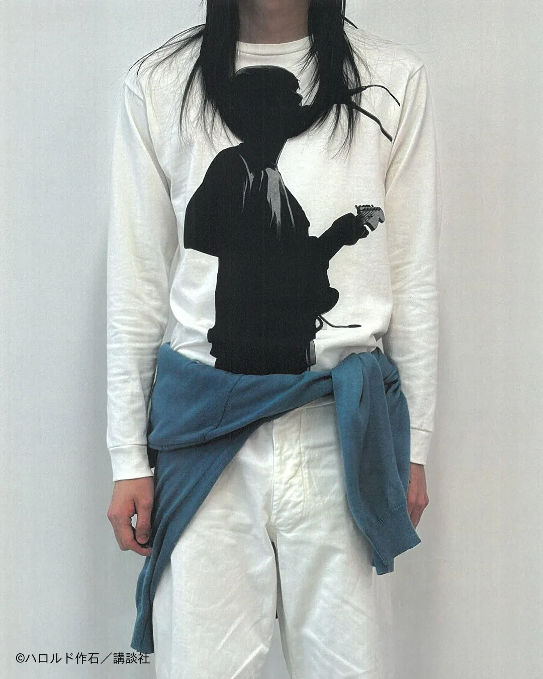 白い長袖Tシャツを着た人物がギターを演奏しているシルエットが描かれている。青いセーターを腰に巻き、白いズボンを履いている。髪は長く、顔は隠れている。