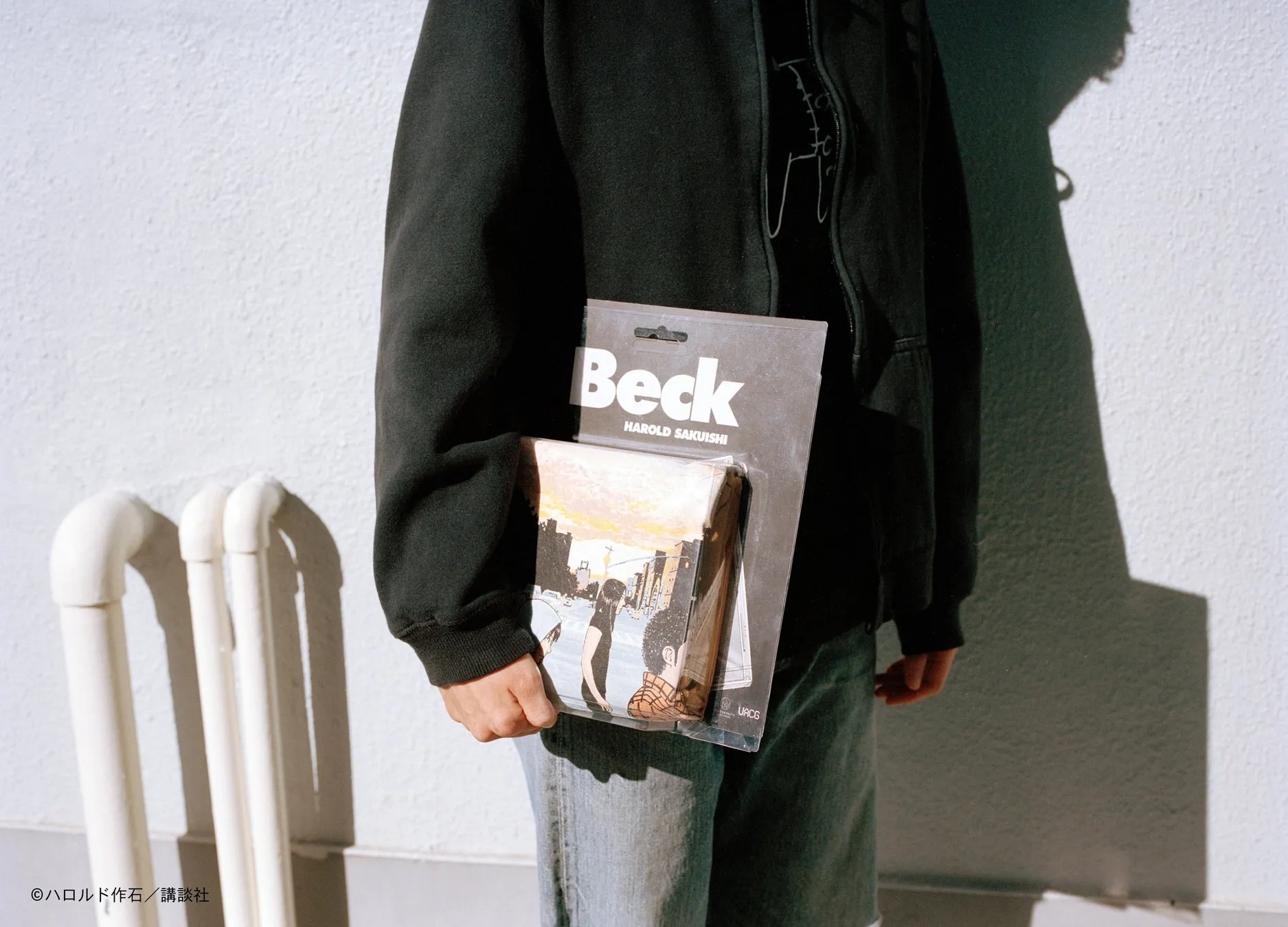 黒いパーカーを着た人物が、漫画「Beck」の単行本を持っている様子を写した写真。背景は白い壁で、日差しが差し込んでいる。