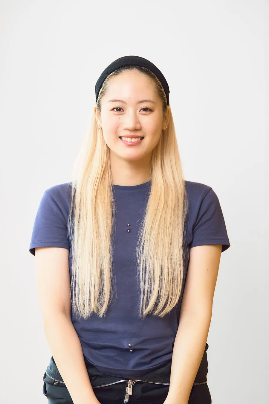 金髪の女性が笑顔でカメラに向かって立っています。彼女は黒いヘッドバンドと濃い色のシャツを着ています。