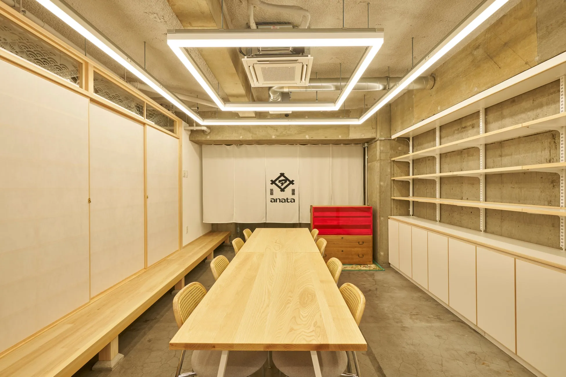 木製のテーブルと椅子が置かれた会議室のような部屋。白いカーテンにはロゴが、壁には棚が設置されている。照明が特徴的。