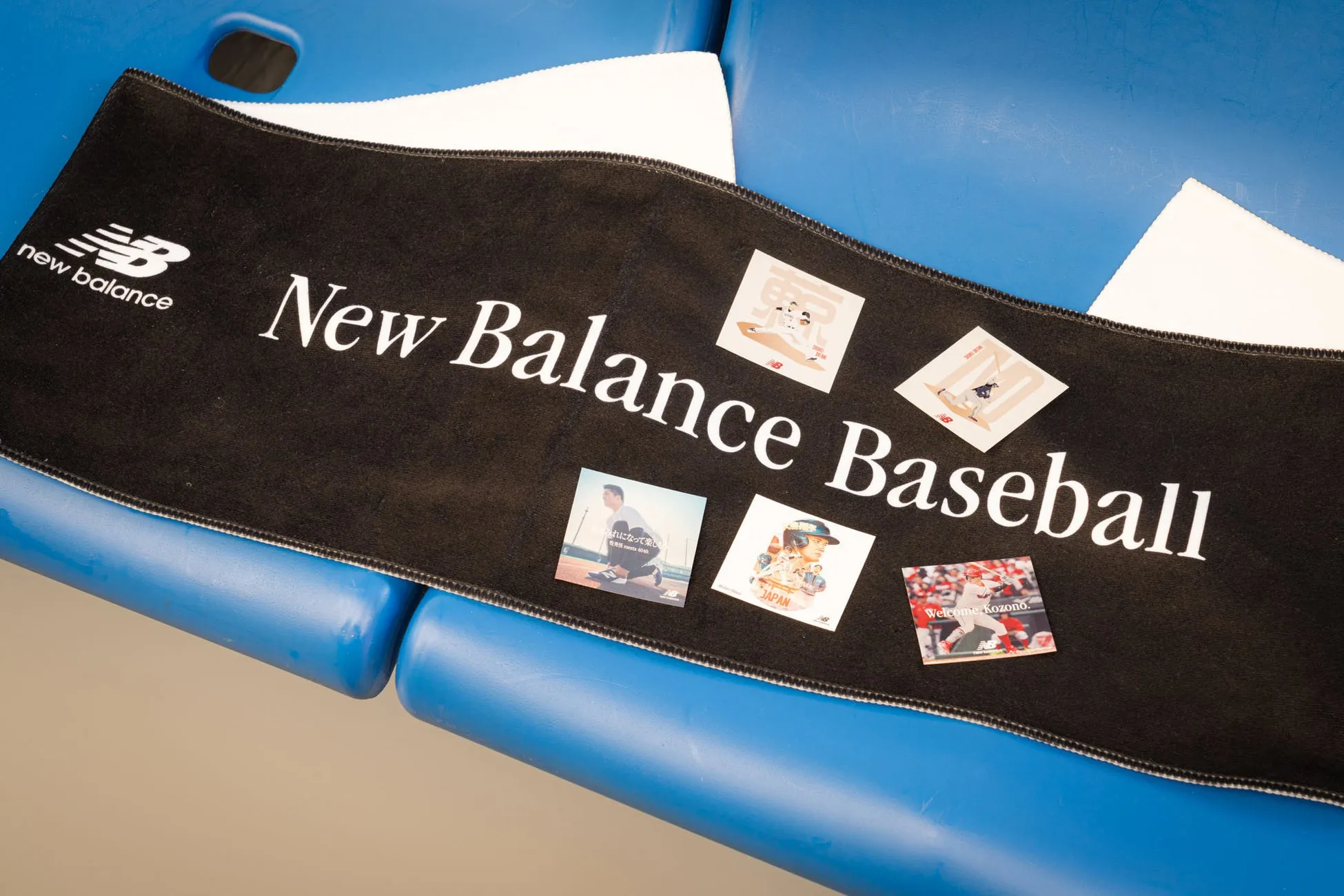 黒いタオルに「New Balance Baseball」の文字とイラストが印刷された画像。タオルは青い椅子の上に置かれており、その上にいくつかのカードが配置されている。