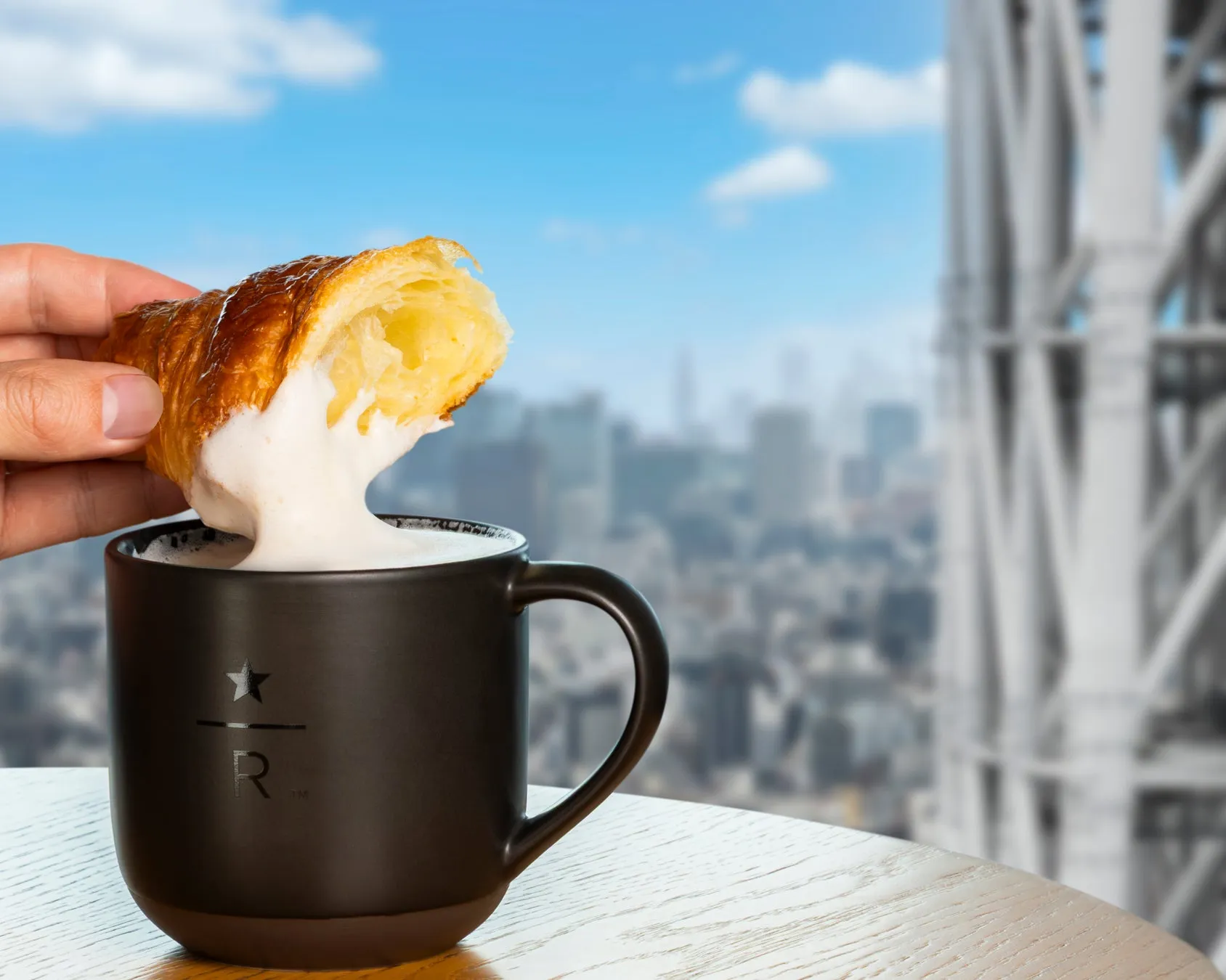 クロワッサンをフォームミルクが入ったマグカップに浸している様子。背景には都会の風景が写っている。
