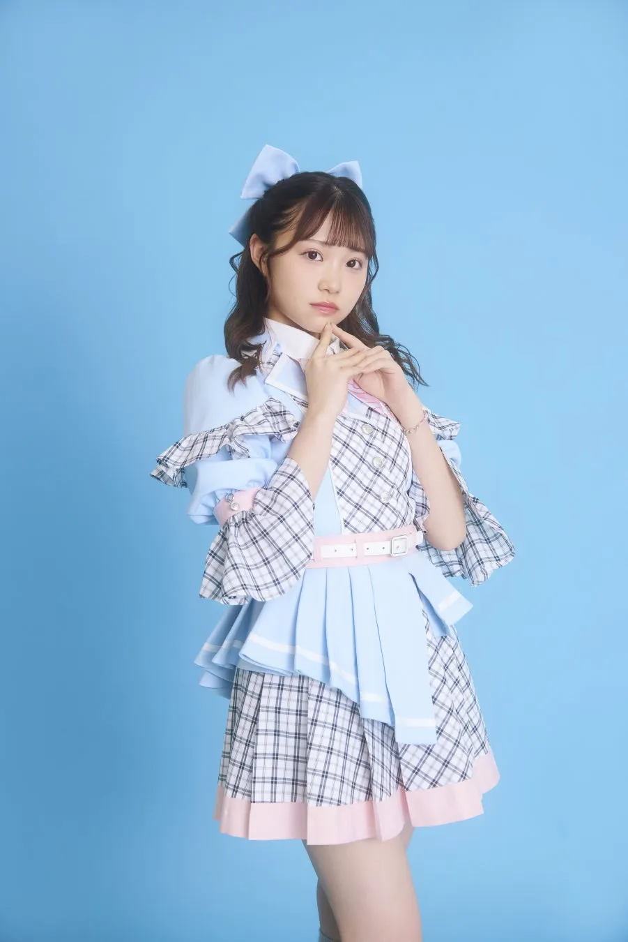 水色の背景に、アイドル風の衣装を着た若い女性が立っている写真です。彼女は青いリボンとフリル付きの衣装を身につけており、可愛らしいポーズをとっています。
