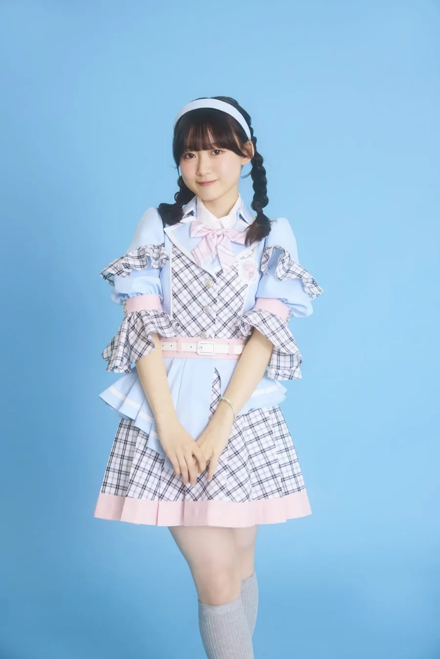 水色の衣装を着た女性のポートレート。彼女は、チェック柄のミニスカート、ピンクのリボン、髪に白いヘッドバンド、そして三つ編みをしている。彼女はカメラを見て微笑んでいる。