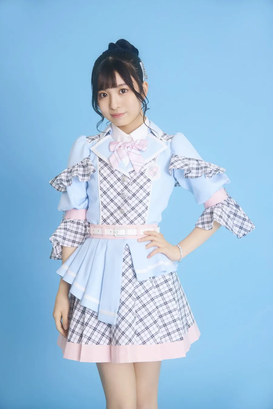 青色の背景の前に立っている若い女性。彼女は青と白のチェック柄の衣装を着ています。彼女は微笑んでいて、カメラを見ています。