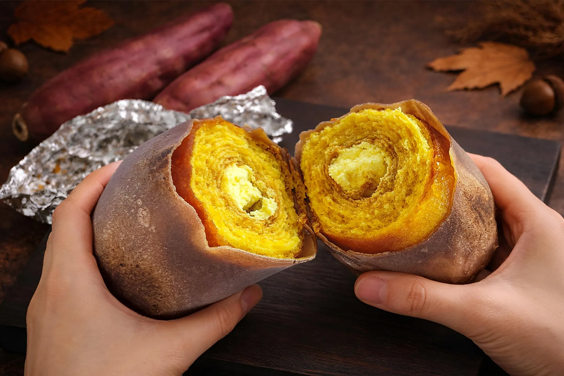 さつまいもの焼き菓子が手で半分に割られている様子。断面は鮮やかな黄色で、層状になっている。背景にはさつまいも、葉、木の実などが見える。